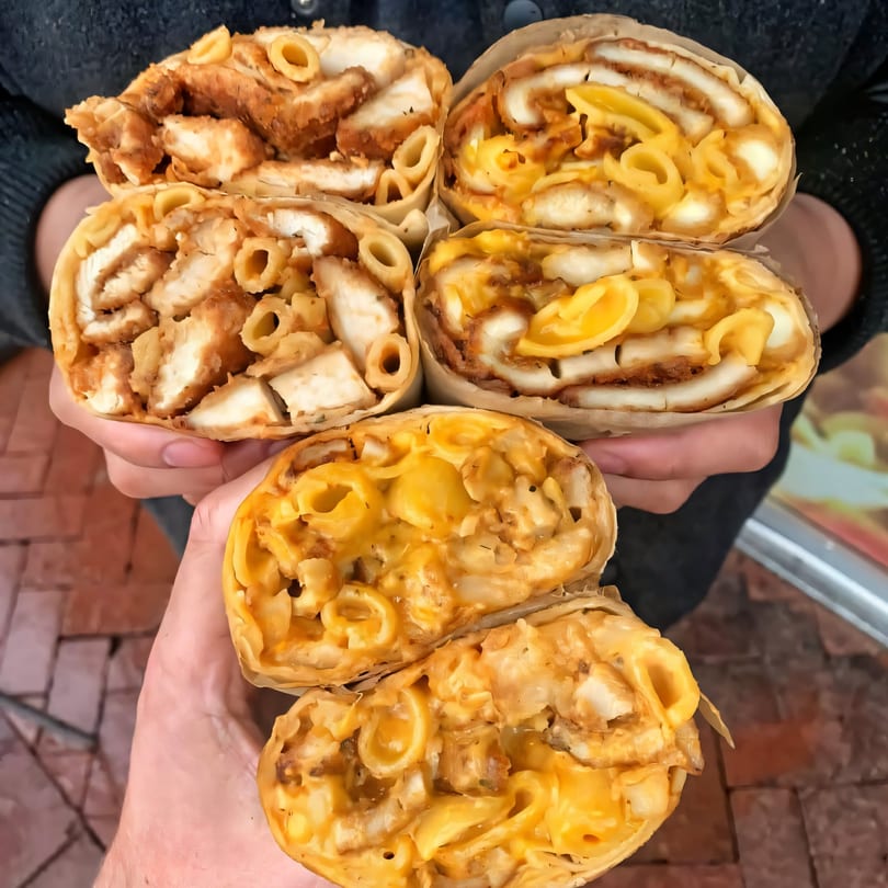 Hands holding five Plainview Bagel Cafe wraps filled with fried chicken and macaroni and cheese.